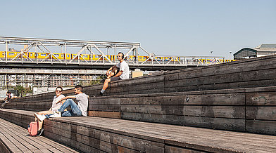 Ein Mann sitzt mit Kopfhörern auf einer Holztribüne im Park am Gleisdreieck, im Hintergrund ist die U-Bahn Hochbahn zu sehen