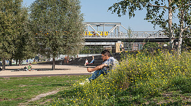 Ein Mann sitzt an einem kleinen Hang im Park am Gleisdreieck und schaut auf sein Handy